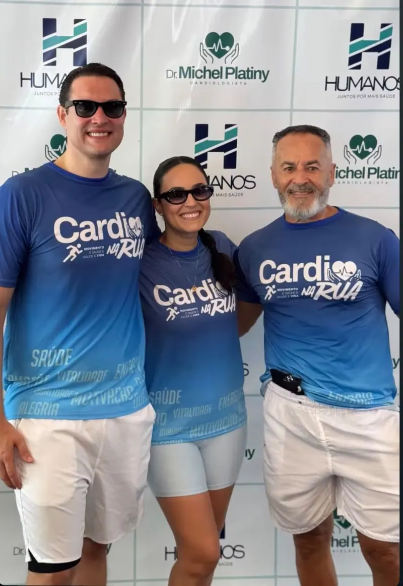 O Dr. Michel e dois participantes de camisa azul do Cardio na Rua em frente ao painel do evento.