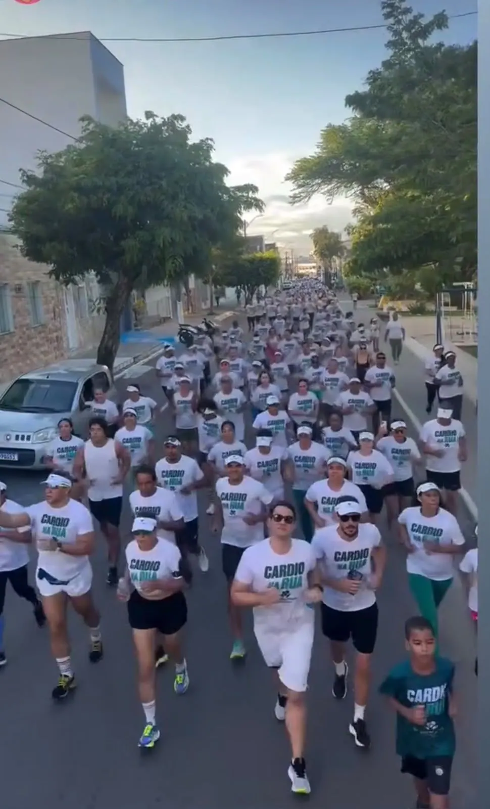 Centenas de participantes de camisa branca do Cardio na Rua correm por uma rua arborizada de Conceição do Coité, com o Dr. Michel à frente.