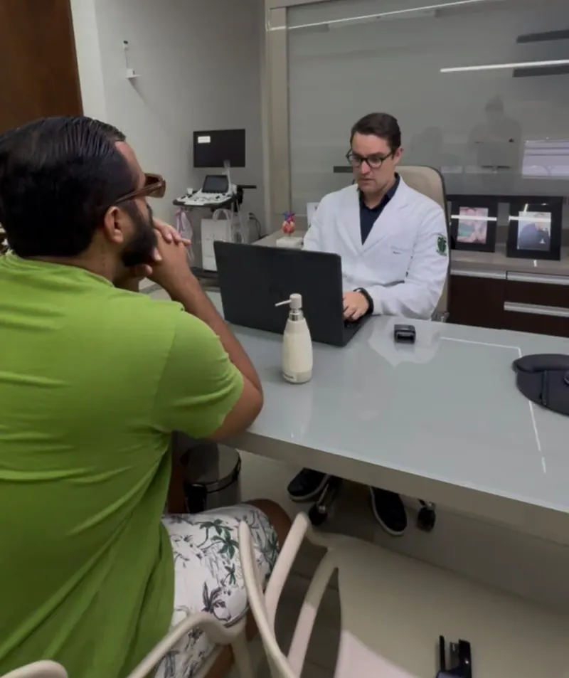 O Dr. Michel, de jaleco, conversa com um paciente durante a consulta no consultório.