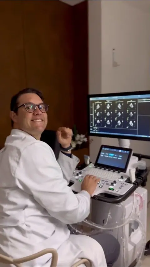 O Dr. Michel, de jaleco, sorri ao lado do aparelho de ecocardiograma.